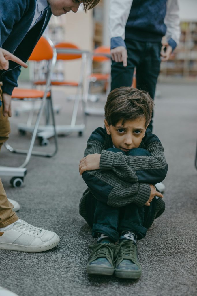 A sad boy sitting on the floor while children point at him in a classroom setting.
