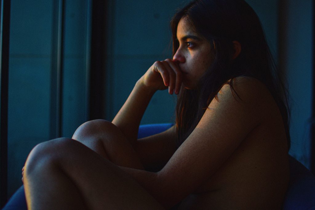 A young woman sits thoughtfully by a window in subdued lighting, evoking introspection.